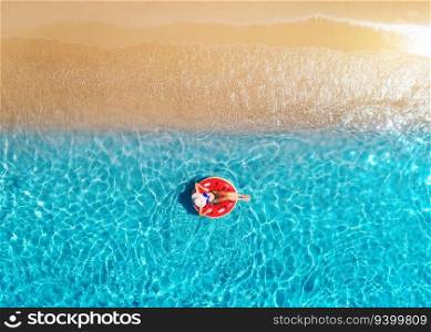 Aerial view of a woman in hat swimming with red swim ring in blue sea at sunrise in summer. Tropical landscape with girl, clear water, waves, sandy beach. Top view. Vacation. Sardinia island, Italy 