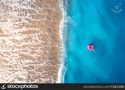 Aerial view of a young woman swimming with pink donut swim ring in clear blue sea with waves at sunset in summer. Tropical landscape with girl, azure water, sandy beach. Top view. Travel and tourism. Aerial view of a young woman swimming with pink donut swim ring
