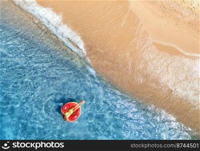 Aerial view of a young woman swimming with red swim ring in blue sea with waves at sunset in summer. Tropical landscape with girl, clear water, sandy beach. Top view. Vacation. Sardinia island, Italy 