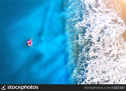 Aerial view of a young woman swimming with the donut swim ring in the clear blue sea with waves at sunset in summer. Tropical landscape with girl, azure water, sandy beach. View from above. Travel. Aerial view of a young woman swimming with the donut swim ring