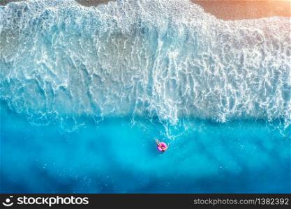Aerial view of a young woman swimming with the donut swim ring in transparent blue sea with waves at sunset in summer. Tropical landscape with girl, azure water, sandy beach. View from above. Travel. Aerial view of a young woman swimming with the donut swim ring