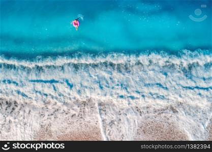 Aerial view of a young woman swimming with the donut swim ring in the clear blue sea with waves at sunset in summer. Tropical landscape with girl, azure water, sandy beach. View from above. Travel. Aerial view of a young woman swimming with the donut swim ring