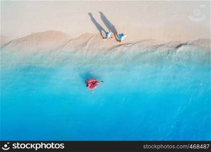 Aerial view of a young woman swimming with the donut swim ring in the blue sea, waves and walking people at sunset in summer. Tropical aerial landscape with girl, azure water, sandy beach. Top view