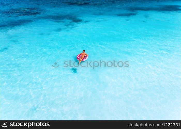 Aerial view of a young woman swimming with the pink donut swim ring in the clear blue sea at sunset in summer. Tropical aerial landscape with girl, clear azure water, sandy beach. Top view. Travel