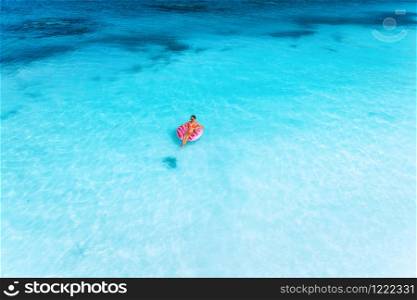 Aerial view of a young woman swimming with the pink donut swim ring in the clear blue sea at sunset in summer. Tropical aerial landscape with girl, clear azure water, sandy beach. Top view. Travel