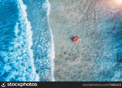 Aerial view of a young woman swimming with the pink donut swim ring in the clear blue sea with beautiful waves at sunset in summer. Tropical aerial landscape with girl, azure water. Top view. Travel