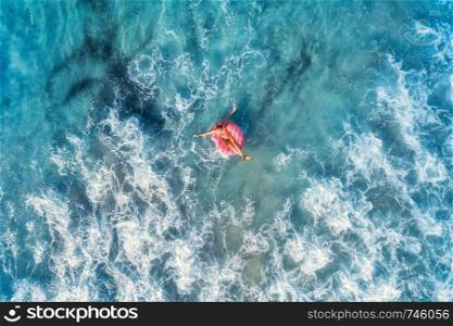 Aerial view of a young woman swimming with the pink donut swim ring in clear blue sea with beautiful waves at sunny day in summer. Tropical aerial landscape with girl, azure water. Top view. Travel