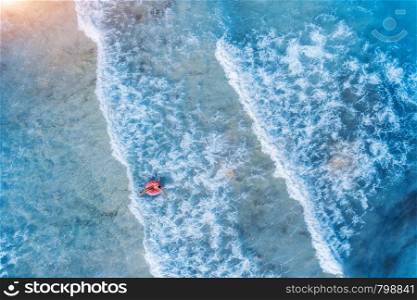 Aerial view of a young woman swimming with the pink donut swim ring in the clear blue sea with beautiful waves at sunset in summer. Tropical aerial landscape with girl, azure water. Top view. Travel. Aerial view of a young woman swimming with the donut swim ring