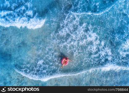 Aerial view of a young woman swimming with the pink donut swim ring in clear blue sea with beautiful waves at sunny day in summer. Tropical aerial landscape with girl, azure water. Top view. Travel. Aerial view of a young woman swimming with the donut swim ring