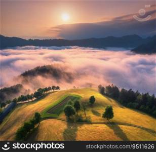 Aerial view of alpine meadows and mountains in pink low clouds at sunrise in autumn. Top drone view of hills with green grass and trees in fog, colorful sky at dawn in fall. Slovenia. Mountain valley 