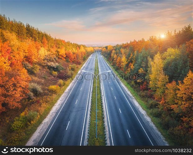 Aerial view of asphalt road in beautiful autumn forest at sunset. Colorful landscape with empty highway, trees with red and orange leaves, blue sky with sun in fall. Top view of roadway. Autumn colors