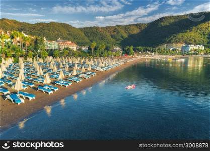 Aerial view of beautiful beach with chaise-lounges and swimming woman on the air mattress in the sea against mountains at sunset. Summer seascape with girl, blue water, sky. Top view. Icmeler, Turkey