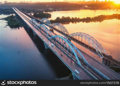 Aerial view of beautiful bridge at sunset in Kiev, Ukraine. Landscape with bridge, river, city, colorful sky with red clouds in summer. Cityscape with road, buildings, reflection in water. Top view