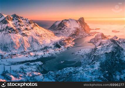 Aerial view of beautiful fjord at sunset in Lofoten Islands, Norway. Winter landscape with snowy mountains, blue sea and sky with golden sunlight. Top view of rocks in snow, village. North coastline