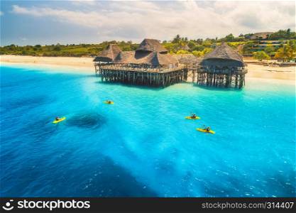 Aerial view of beautiful hotel and people in kayaks at sunset in summer. Zanzibar, Africa. Top view. Landscape with wooden hotel on the sea, azure water, sandy beach, green trees, canoe. Luxury resort