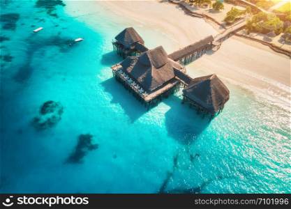 Aerial view of beautiful hotel on the water in ocean at sunset in summer. Zanzibar, Africa. Top view. Landscape with wooden hotel on the sea, azure water, sandy beach, green palm trees. Luxury resort