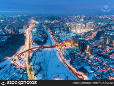 Aerial view of beautiful modern city at cold night in winter. Top view of traffic on roads, buildings, snowy streets with illumination. Skyline. Cityscape from drone. Aerial view of highway, downtown