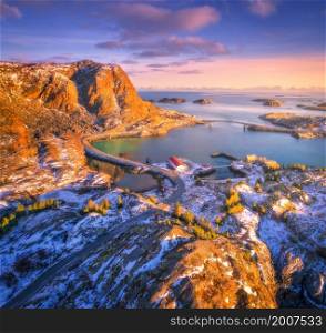 Aerial view of beautiful mountains, bridge, small islands in the sea, road, purple sky at sunset. Lofoten islands, Norway in winter. Top view of road, snowy rocks, stones, sea coast, water. Top view