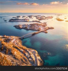 Aerial view of beautiful mountains, bridge, small islands in the sea, road, orange sky at sunset. Lofoten islands, Norway in winter. Top view of road, snowy rocks, stones, sea coast, water. Top view