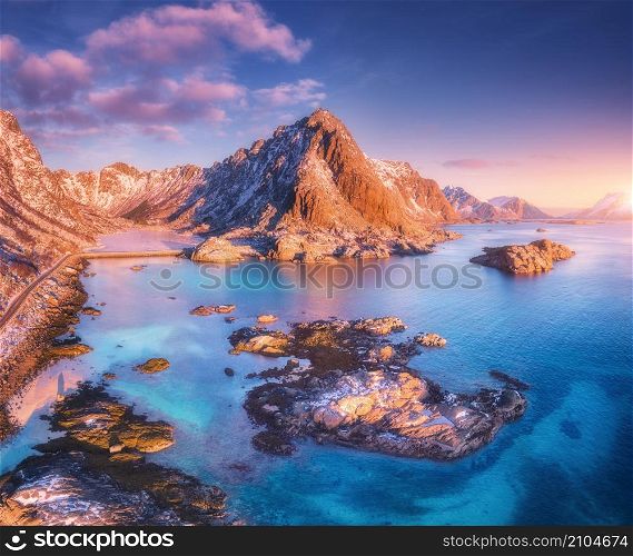 Aerial view of beautiful mountains, small islands in the sea, road, blue sky at sunset in Lofoten islands, Norway in winter. Top view of road, snowy rocks, stones, sea coast, clear water. Top view