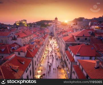 Aerial view of beautiful old city at sunset. Top view of houses with red roofs, city lights, historical centre, architecture, walking people in illuminated streets at night in Dubrovnik, Croatia