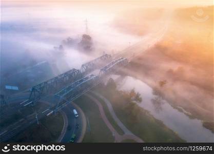 Aerial view of beautiful railroad bridge and river in fog at sunrise in fall. Autumn landscape with foggy meadows, mist over the water, trees, railway station, orange sky with gold sunlight. Top view