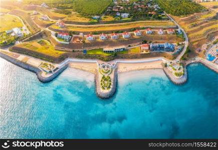 Aerial view of beautiful sandy beach, blue sea, promenade, houses, pool, umbrellas, water, green trees on the hills at sunset in summer. Top view of seafront and cottage town. Tropical landscape.