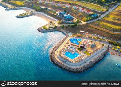 Aerial view of beautiful sandy beach, blue sea, restaurants on the promenade, pool, umbrellas, clear water, cottage town, green trees at sunset in summer. Top view of seafront. Tropical landscape