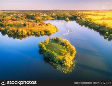 Aerial view of beautiful small island with green trees and boats on the river at sunset in summer. Colorful landscape with island, meadow, forest and sky reflection in blue water. Top view. Nature . Aerial view of beautiful small island with green trees and boats