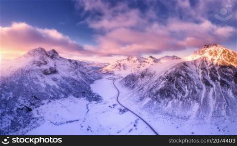 Aerial view of beautiful snowy mountains in winter at sunset. Lofoten Islands, Norway. Colorful landscape with road, rocks in snow, violet sky with clouds. Top view from drone. Nature. View from above