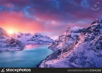 Aerial view of beautiful snowy mountains, sea, road, sky with pink clouds at sunset in winter. Lofoten islands, Norway. Top view of rocks in snow, sea coast, azure water. View from above. Nature