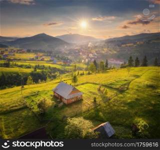 Aerial view of beautiful village in Carpathian mountains at sunset in summer. Colorful landscape with green meadows, houses with gardens, trees, sky with clouds. Top view of mountain countryside