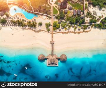 Aerial view of beautiful wooden hotel in Indian ocean at sunset in summer. Zanzibar, Africa. Top view. Tropical landscape with building on the sea, clear blue water, sandy beach, palm trees and pool