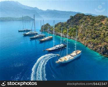 Aerial view of beautiful yachts and boats on the sea at sunset in summer. Gemiler Island in Turkey. Top view of luxury yachts, sailboats, clear blue water, beach, mountain and green forest. Travel