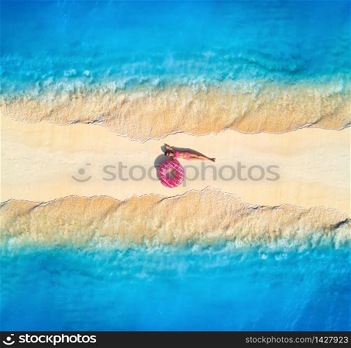 Aerial view of beautiful young lying woman with pink donut swim ring on the white sandy beach and sea with waves on the both sides at sunset. Summer travel. Top view of slim girl, blue water, sandbank. Aerial view of woman on sandy beach with waves on the both sides