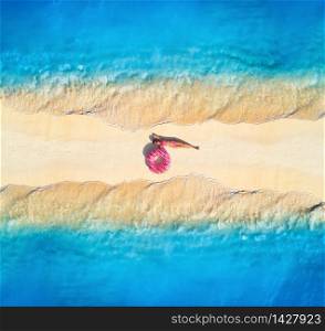 Aerial view of beautiful young lying woman with pink donut swim ring on the white sandy beach and sea with waves on the both sides at sunset. Summer travel. Top view of slim girl, blue water, sandbank. Aerial view of woman on sandy beach with waves on the both sides