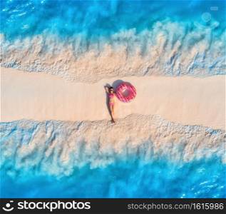 Aerial view of beautiful young lying woman with pink donut swim ring on the white sandy beach and sea with waves on the both sides at sunset. Summer travel. Top view of slim girl, blue water, sandbank