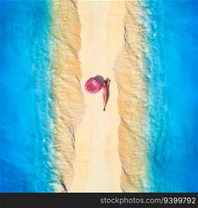 Aerial view of beautiful young lying woman with pink donut swim ring on the white sandy beach and sea with waves on the both sides at sunset. Summer travel. Top view of slim girl, blue water, sandbank