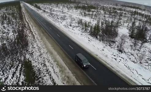 Aerial view of black minivan driving along winter road. North territory with wide open spaces and thin-sown trees