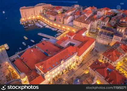Aerial view of boats and yachts and beautiful architecture at night in Dubrovnik, Croatia. Top view of old city at sunset. Port in the sea, city lights, historical centre, buildings and red roofs