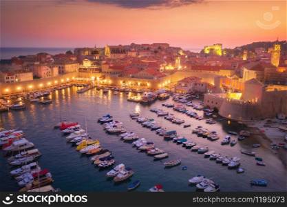 Aerial view of boats and yachts and beautiful architecture at night in Dubrovnik, Croatia. Top view of old city at sunset. Port in the sea, city lights, historical centre, buildings and colorful sky