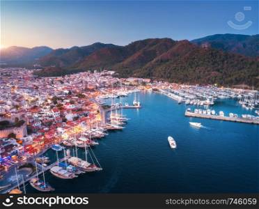 Aerial view of boats and yachts and beautiful architecture at sunset in Marmaris, Turkey. Landscape with boats, sea, houses, city illumintaion, mountains. Top view of harbor and blue sky at dusk