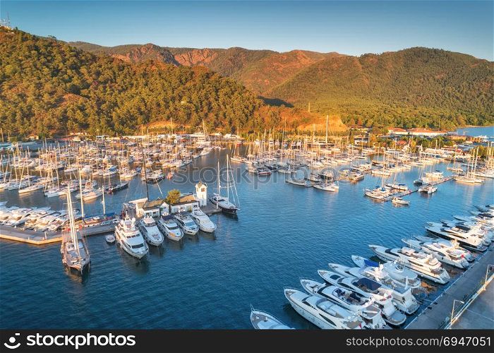 Aerial view of boats and yachts at sunset in Turkey. Colorful landscape with boats in marina bay, sea, mountains, forest, blue sky. Top view from drone of harbor with luxury yacht, sailboat. Travel. Aerial view of boats and yachts at sunset