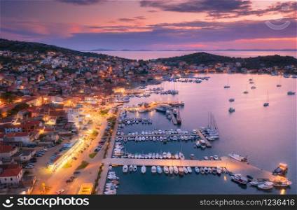Aerial view of boats and yachts in port and city at night. Summer landscape with city lights, buildings, illuminated streets, mountain, motorboats, blue sea, purple sky at sunset. Top view. Croatia