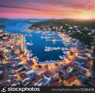Aerial view of boats and yachts in port and city at sunset. Summer landscape with city lights, buildings, illuminated streets, mountain, motorboats, blue sea, red sky at night. Top view of Croatia