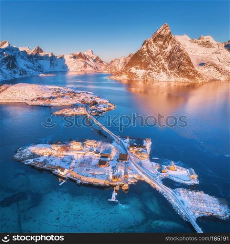 Aerial view of bridge over the sea and snowy mountains in Lofoten Islands, Norway. Reine and Hamnoy at sunrise in winter. landscape with blue water, rocks, buildings, rorbuer, road, bridge. Top view