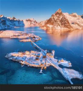 Aerial view of bridge over the sea and snowy mountains in Lofoten Islands, Norway. Reine and Hamnoy at sunrise in winter. landscape with blue water, rocks, buildings, rorbuer, road, bridge. Top view