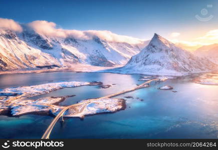 Aerial view of bridge over the sea and snowy mountains in Lofoten Islands, Norway. Fredvang bridges at sunset in winter. Landscape with blue water, rocks in snow, road and sky with cloads. Top view