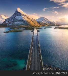 Aerial view of bridge over the sea and snowy mountains in Lofoten Islands, Norway. Fredvang bridges at sunset in winter. Landscape with blue water, rocks in snow, road, sky with clouds. Top drone view