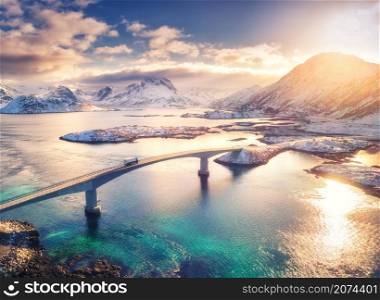 Aerial view of bridge, sea and snowy mountains in Lofoten Islands, Norway. Fredvang bridges at sunset in winter. Landscape with blue water, rocks in snow, road and sky with clouds. Top view from drone